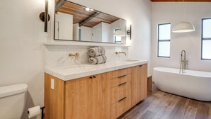 Une salle de bain moderne avec un meuble-lavabo en bois, un grand miroir, une baignoire sur pieds et une fenêtre laissant entrer la lumière naturelle.