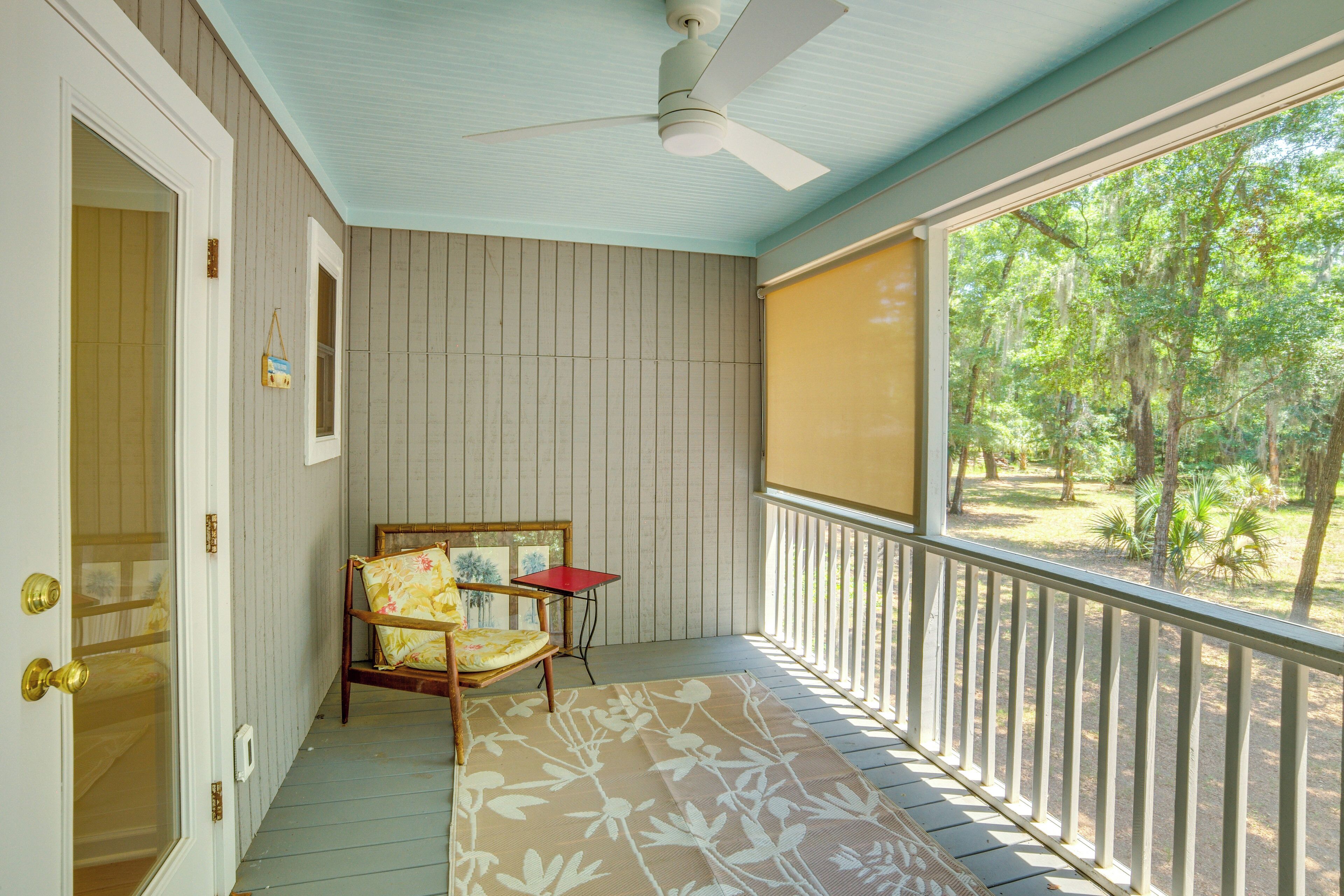 A covered porch with a ceiling fan, a wooden chair with a floral cushion, a small table, and a view of trees through a screened window.