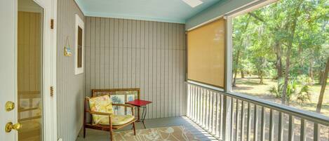 A covered porch with a ceiling fan, a wooden chair with a floral cushion, a small table, and a view of trees through a screened window.