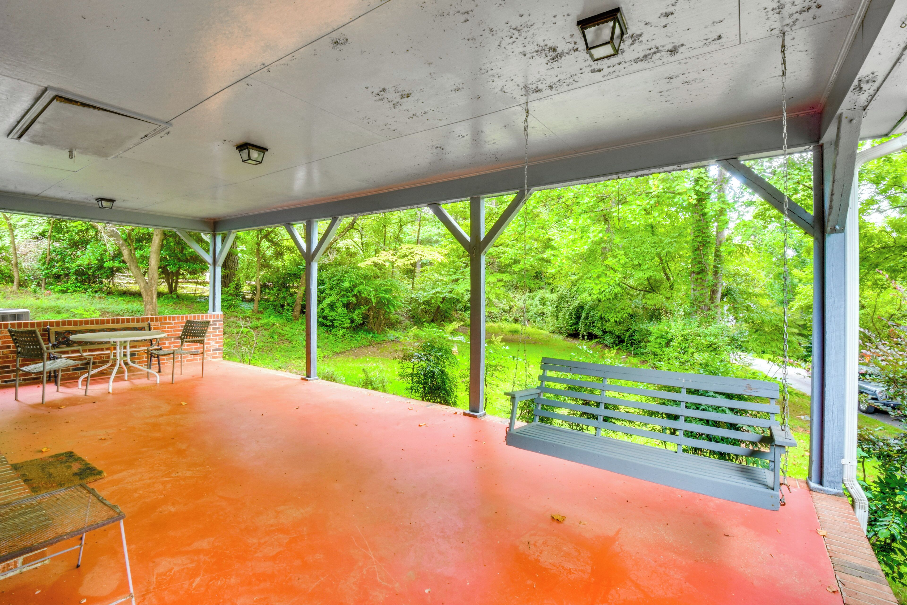 A covered outdoor area with a table and chairs, a bench, and a view of a green forest.