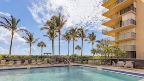 A swimming pool with clear blue water, surrounded by lounge chairs and palm trees, with a building in the background. - Oceanfront Fort Pierce Condo: Pool & Beach Access! (Fort Pierce)