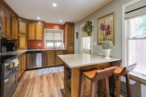 A kitchen with wooden cabinets, a stainless steel dishwasher, and a bar area with stools. - Mt Dora Garden Cottage w/ Patios, Walk to Downtown (Mount Dora)