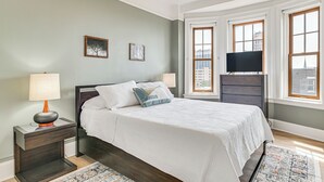 Une chambre à coucher avec un lit, des tables de chevet, une lampe, un téléviseur et de grandes fenêtres donnant sur la ville.