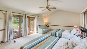 Habitación de hotel con dos camas, ventilador de techo, puerta corrediza de vidrio al exterior y un pequeño rincón de descanso con una planta.