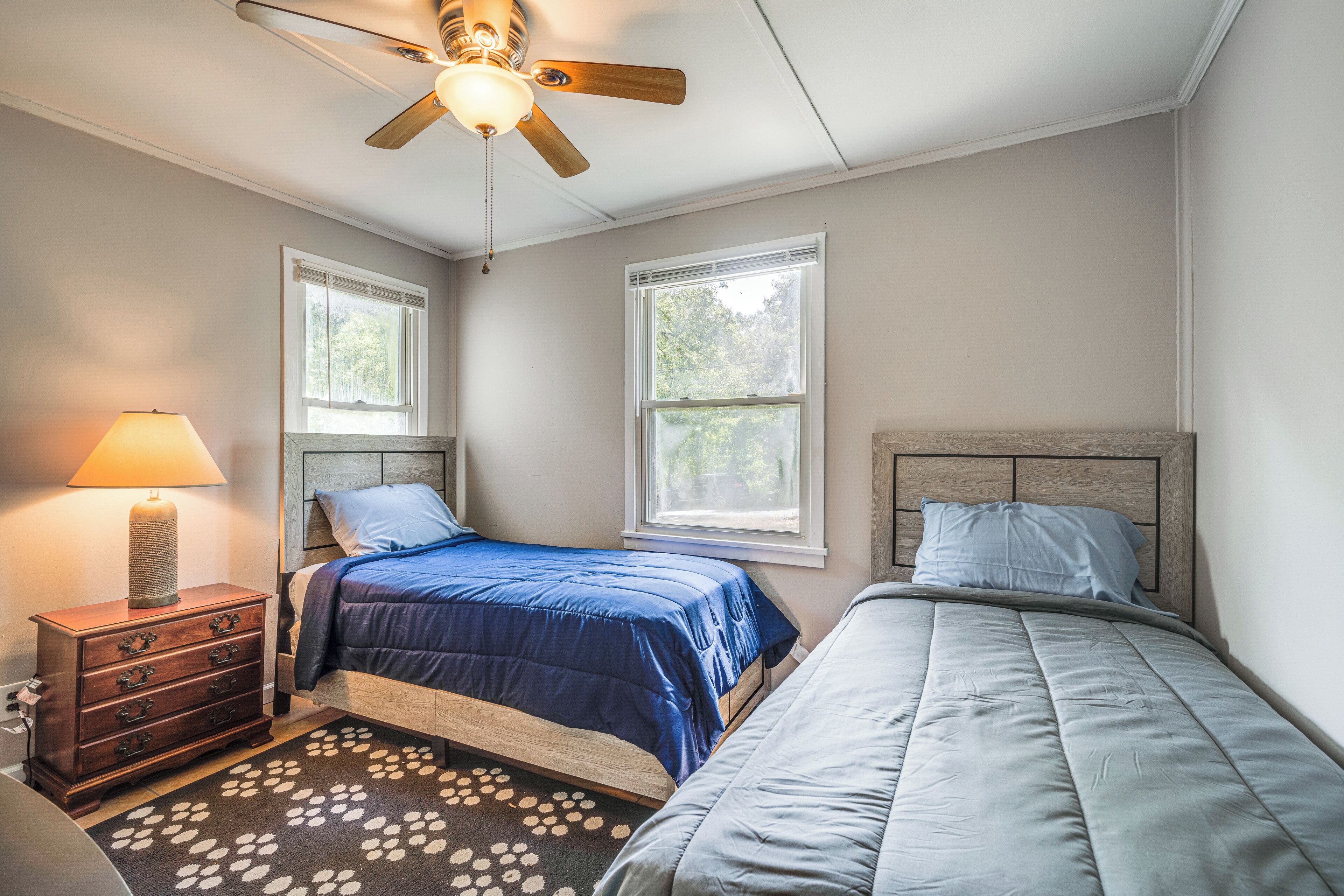 A bedroom with two beds, a ceiling fan, a nightstand with a lamp, and a window with blinds.