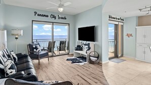 A living room with a large window overlooking the ocean, a ceiling fan, a flat-screen TV, and a sofa with patterned cushions. - Origin 1203/1205 - Three Bedroom Connector Unit - Sunset Gulf View (Panama City Beach)