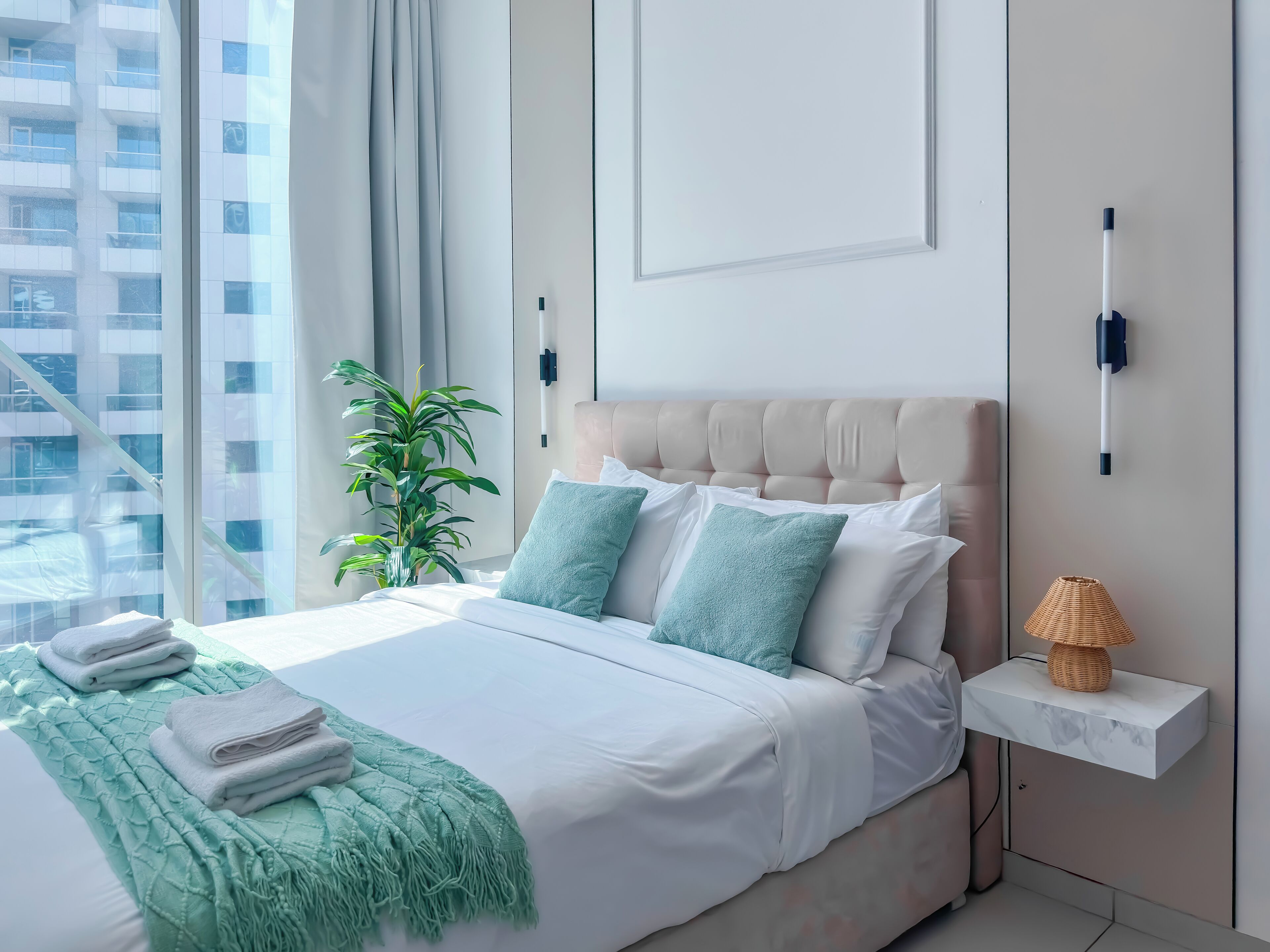 Ein modernes Schlafzimmer mit einem großen Bett, einem Nachttisch mit Lampe und Blick auf eine Stadtlandschaft durch das Fenster.