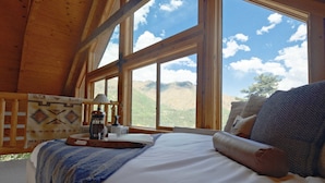 A bedroom with a large window, a bed with a blue blanket, a wooden headboard, and a view of mountains. - Hillside Cabin - Mt Views - Patio Decks - Hot Tub (Cascade)