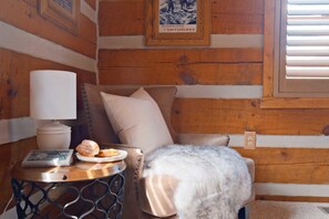 A cozy cabin interior with a wooden wall, a bed with a fluffy blanket, a small table with a lamp, and a framed picture on the wall.