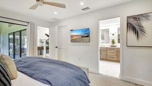 Une chambre à coucher avec un lit, un ventilateur de plafond, un tableau, une salle de bain avec un miroir et une porte donnant sur une autre pièce.