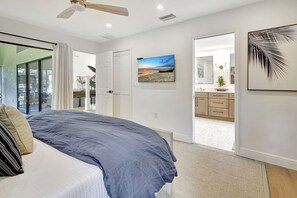 A bedroom with a bed, a ceiling fan, a painting, a bathroom with a mirror, and a door leading to another room.