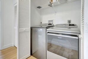 A laundry room with a front-loading washer and dryer, a vent hood, and a white door with louvered panels. - Luxury, Privacy and Close to Beach (Bonita Springs)