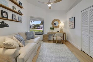 A well-lit room with a beige sofa, a desk with a chair, a round mirror, and a window with blinds.