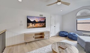 A modern living room with a flat-screen TV mounted on the wall, a wooden entertainment unit, a ceiling fan, and a large window with a view of a brick building.