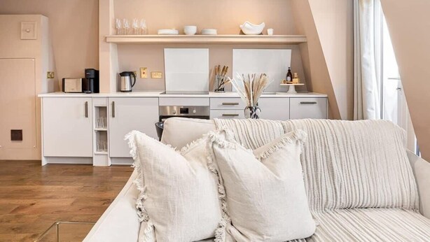 A modern kitchen with white cabinetry, a built-in oven, and a coffee maker. There is a white sofa with ruffled pillows in the foreground. - St Johns Wood Terrace Retreat (London)