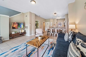 Una sala de estar con un sofá, una mesa de centro y un televisor. Hay un comedor con una mesa y sillas. La habitación tiene una paleta de colores neutros.