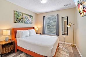 A hotel room with a bed, a nightstand with a lamp, a window with blinds, a painting on the wall, and a floor lamp. - Amazing 3BR Home w Balcony very close to Disney! (Kissimmee)
