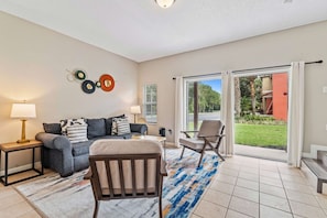 A modern living room with a sofa, armchairs, a coffee table, and a rug. There is a large window with a view of greenery outside. - Amazing 3BR Home w Balcony very close to Disney! (Kissimmee)