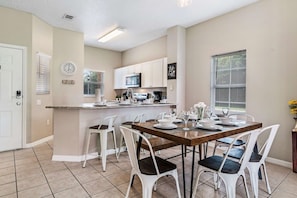 A modern kitchen with white cabinets, a microwave, and a dining area with a wooden table and white chairs. - Amazing 3BR Home w Balcony very close to Disney! (Kissimmee)
