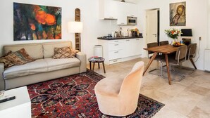 Una sala de estar con sofá, una mesa de centro, alfombra, zona de comedor con mesa y sillas, cocina con armarios y electrodomésticos, y obras de arte en las paredes.