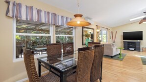 A dining area with a glass-top table and wicker chairs, a large window with curtains, and a television in the background. - Magic Sands Light Hawaii Oasis: Steps to Beach, AC (Kailua Kona)