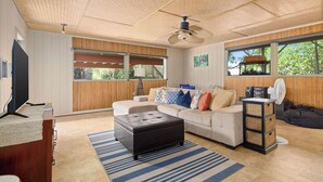A modern living room with a sectional sofa, a black ottoman, a wooden ceiling, and large windows with a view of greenery. - Magic Sands Light Hawaii Oasis: Steps to Beach, AC (Kailua Kona)