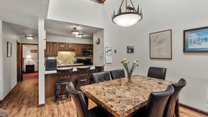 A dining area with a large wooden table and chairs, a chandelier, and a kitchen in the background. - Cozy Mountain Retreat at Timber Run 101! (Steamboat Springs)