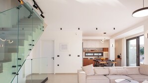 Un interior moderno con una escalera de cristal, una sala de estar con sofá y mesa, y una cocina al fondo.