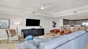 Ein modernes Wohnzimmer mit einem großen Sofa, einem Couchtisch und einem Fernseher. Es gibt eine Küchenzeile mit einer Mikrowelle und einem Kühlschrank.