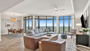 Ein großzügiger Wohnraum mit einer großen, grauen Eckcouch, einem Couchtisch und zwei Sesseln. Es gibt einen Essbereich mit einem Holztisch und Stühlen sowie einen Balkon mit Landschaftsblick.