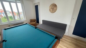 Quarto com mesa de bilhar, sofá, cadeira e vista para edifícios por grandes janelas.