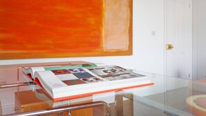 A room with a glass table displaying a book and a magazine. A large orange painting is on the wall. - Fulham Road X by Onefinestay (London)