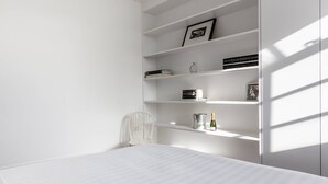 A minimalist bedroom with a bed, white walls, and built-in shelving with books and decorative items. - Fulham Road X by Onefinestay (London)