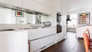 A modern kitchen with white cabinetry, a built-in oven, and a toaster. There is a dining area with a wooden chair and a table. - Fulham Road X by Onefinestay (London)
