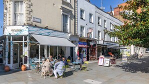 An outdoor cafe with tables and chairs, a sign for a money exchange, and a barber shop. - St Stephen's Gardens VII by Onefinestay (London)
