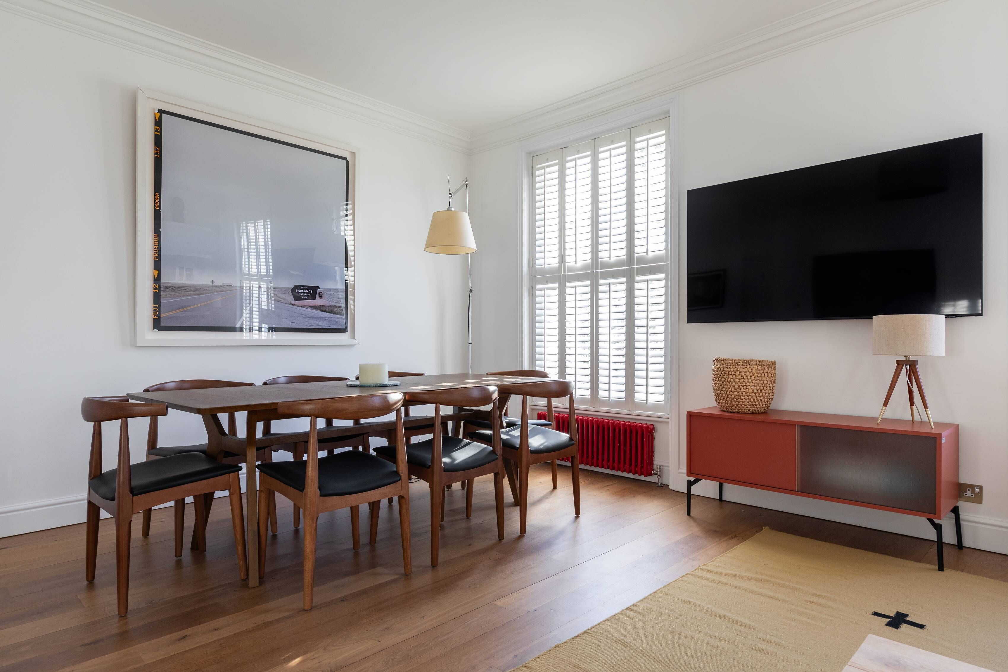 A dining area with a wooden table and chairs, a large framed picture on the wall, a television mounted above a red cabinet, and a window with blinds.