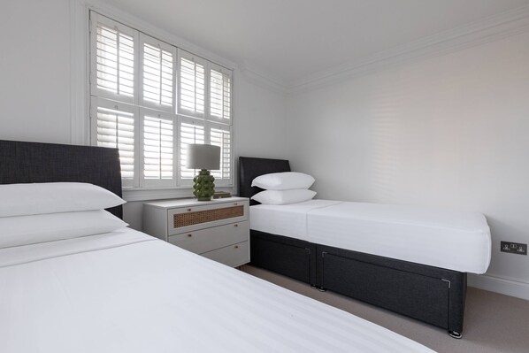 A bedroom with two beds, a nightstand with a lamp, and a window with blinds. - St Stephen's Gardens VII by Onefinestay (London)