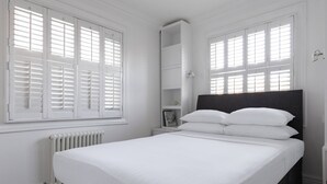 A white bedroom with a bed, a nightstand, a built-in shelf, and a radiator. - St Stephen's Gardens VII by Onefinestay (London)