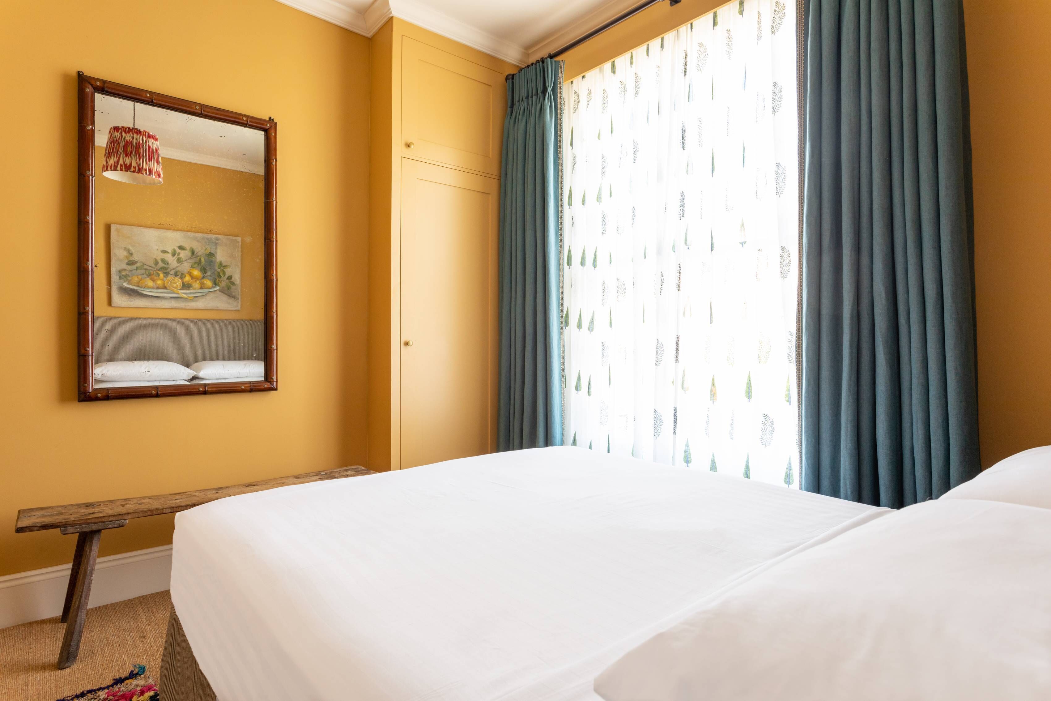 Una habitación de hotel con una cama, un banco de madera, un cuadro y una ventana con cortinas.