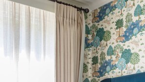 Een slaapkamer met een bed, gordijnen, een hoofdbord en een muur met behang met een natuurthema.
