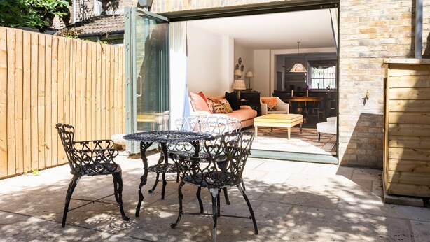 Een terras buiten met een tafel en stoelen, een houten hek en uitzicht op een kamer met een bank en lampen.