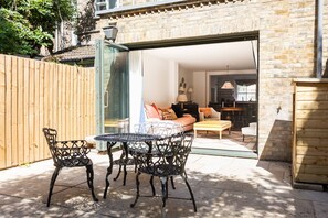 An outdoor patio with a table and chairs, a wooden fence, and a view into a room with a sofa and lamps. - Vernon Street by Onefinestay (London)