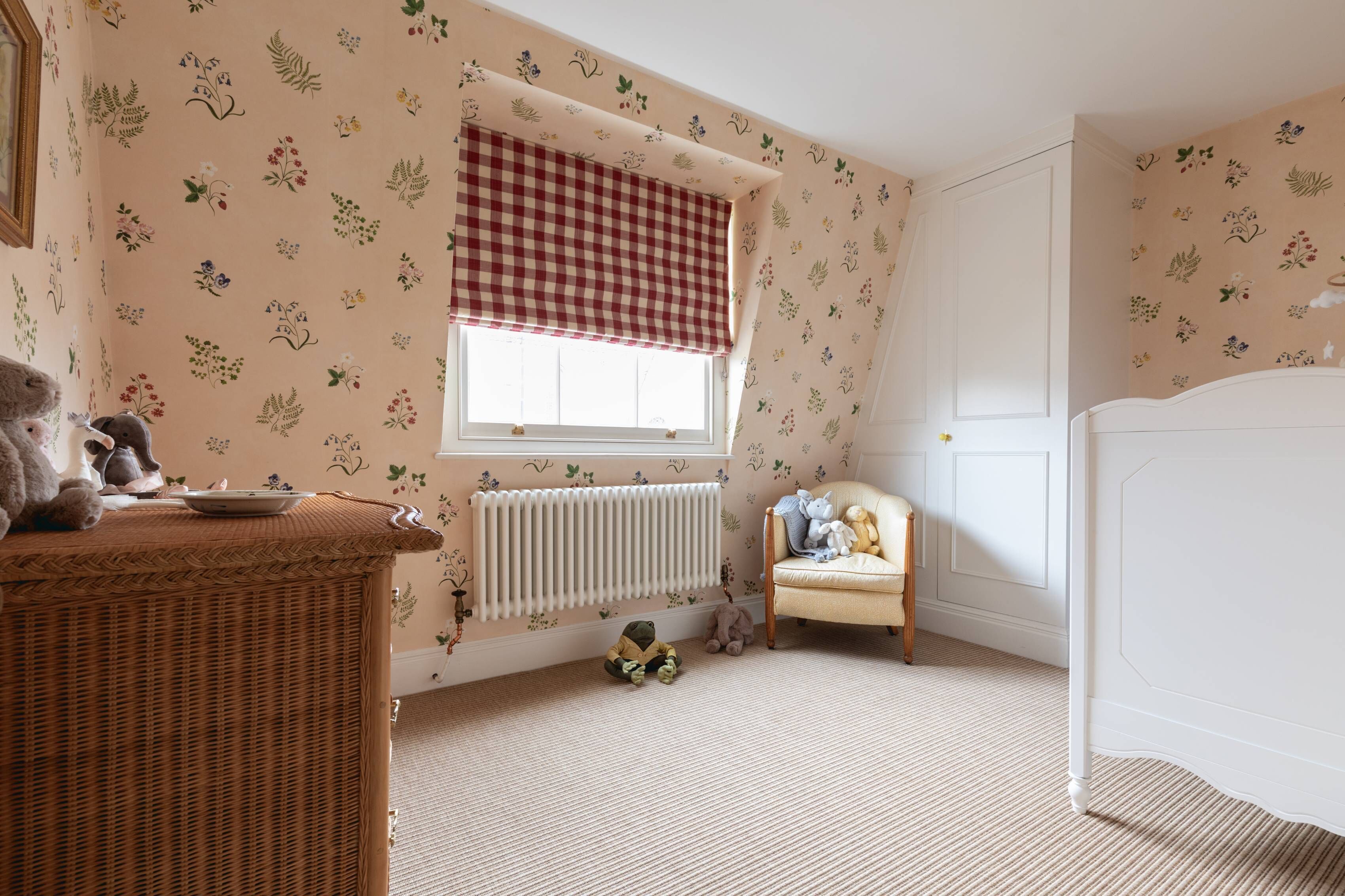 A child's room with floral wallpaper, a wicker side table, a radiator, a window with a checkered blind, a chair, and a bed.