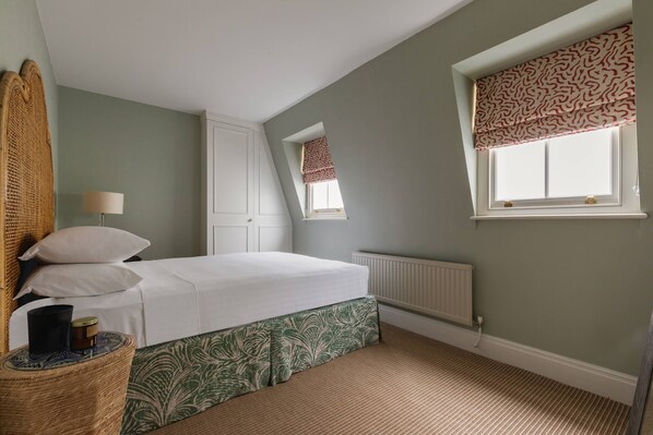 A bedroom with a bed, a radiator, a window with a patterned blind, and a small window with a red and white blind.