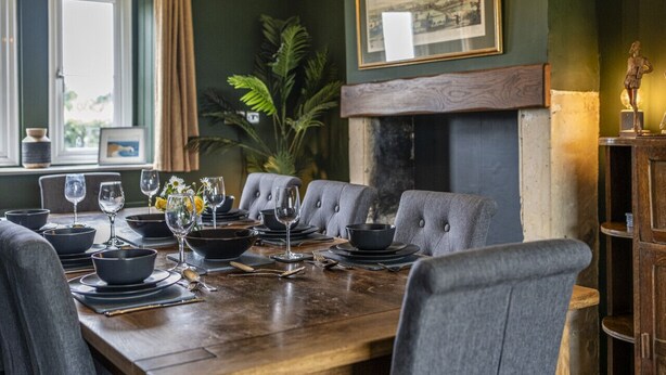 A dining room with a long wooden table set for six, upholstered chairs, a fireplace, and a framed picture on the wall. - Gorgeous Rural 4 Bed Farm Cottage - With Hot Tub (Norton Saint Philip)