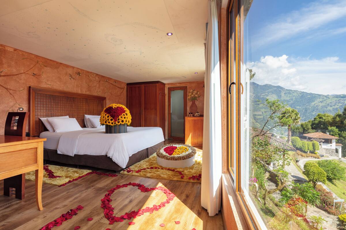 A bedroom with a bed, a wooden nightstand, a vase with flowers, and a view of a mountainous landscape.