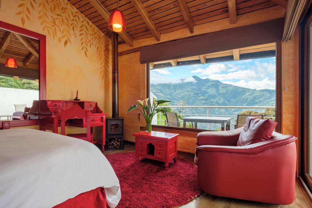 A bedroom with a large bed, a red sofa, a small red desk, a red rug, and a view of mountains through a large window.