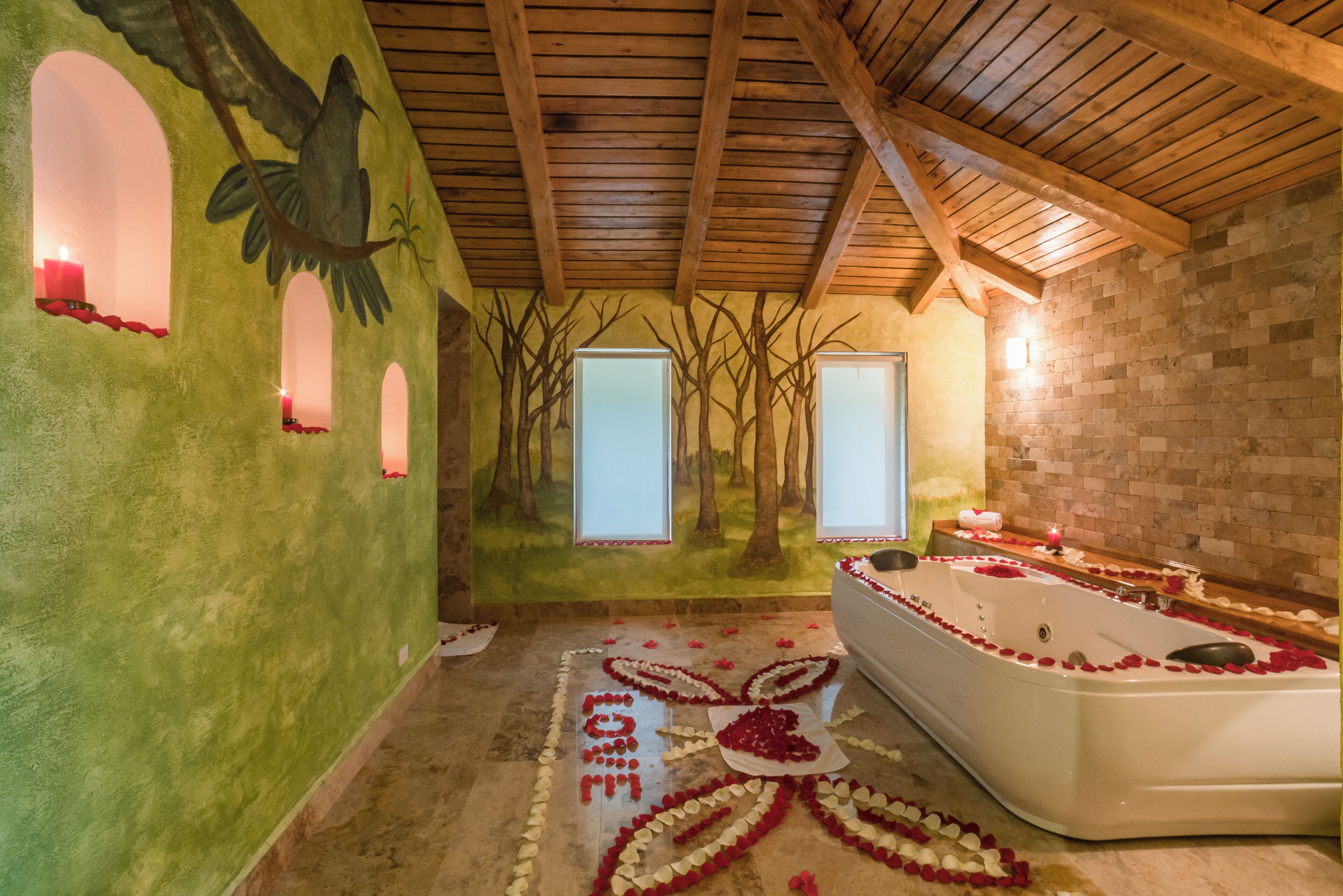 A spa room with a large bathtub surrounded by rose petals, a mural of trees, and a wooden ceiling.