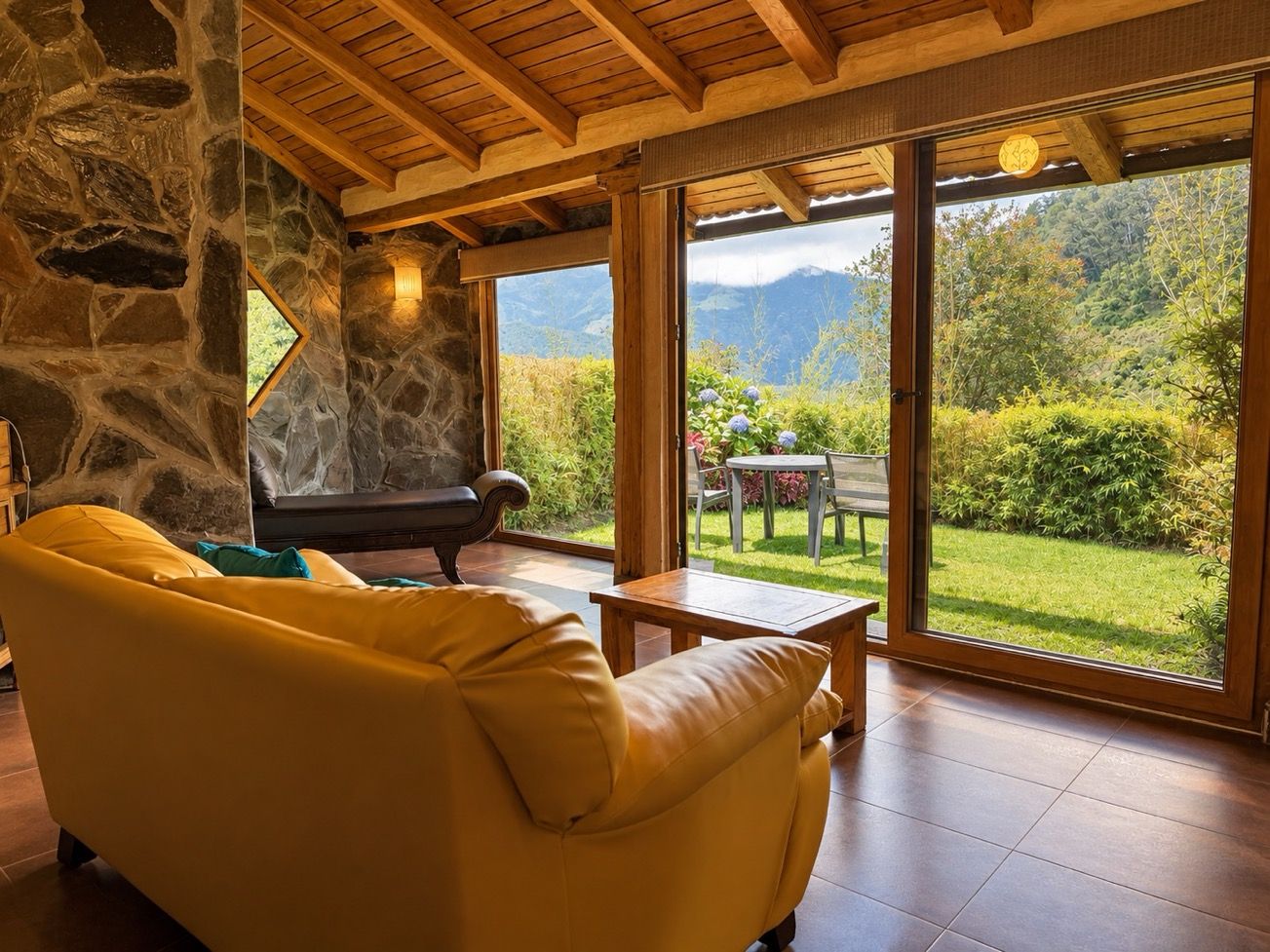 Una habitación con pared de piedra, techo de madera, un sofá amarillo, una mesa y vistas a un jardín y montañas.