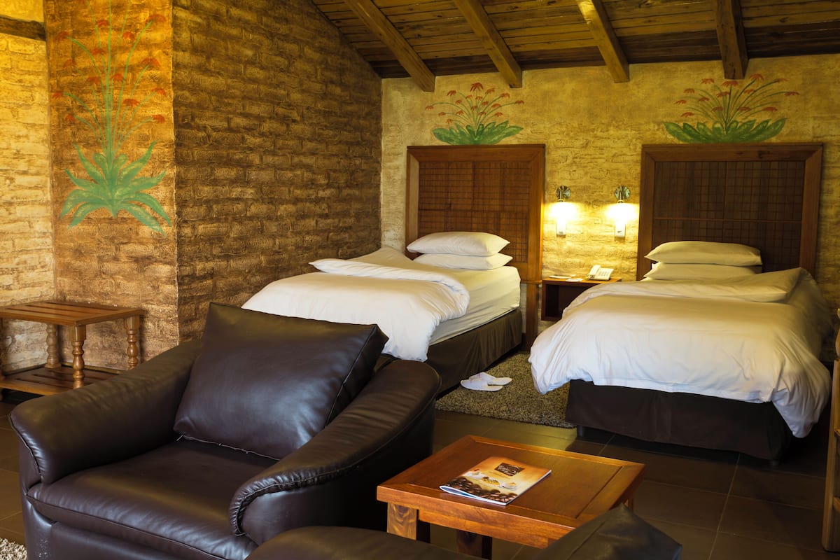 A hotel room with two beds, a leather sofa, a wooden coffee table, and a brick wall with floral artwork.